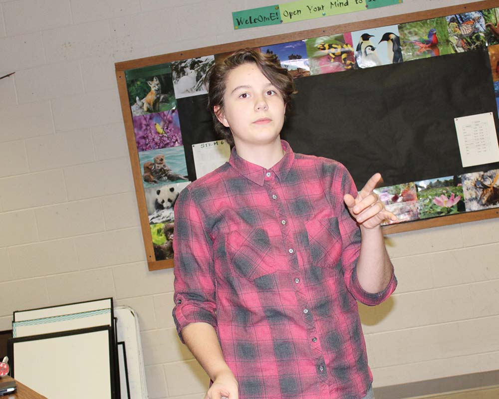 student standing in classroom pointing across room to other students