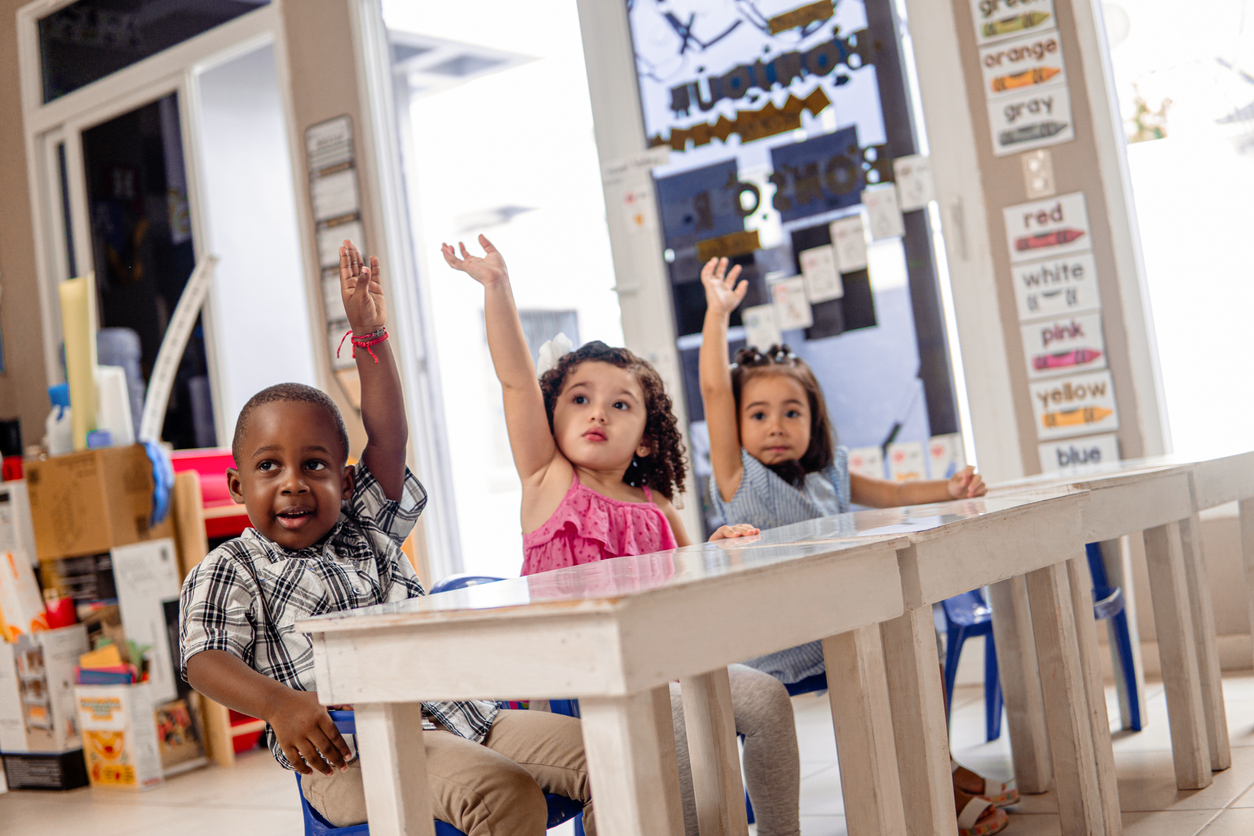 Three Little Children With Your Hands Up In A Class. Children raising hands at desk in classroom